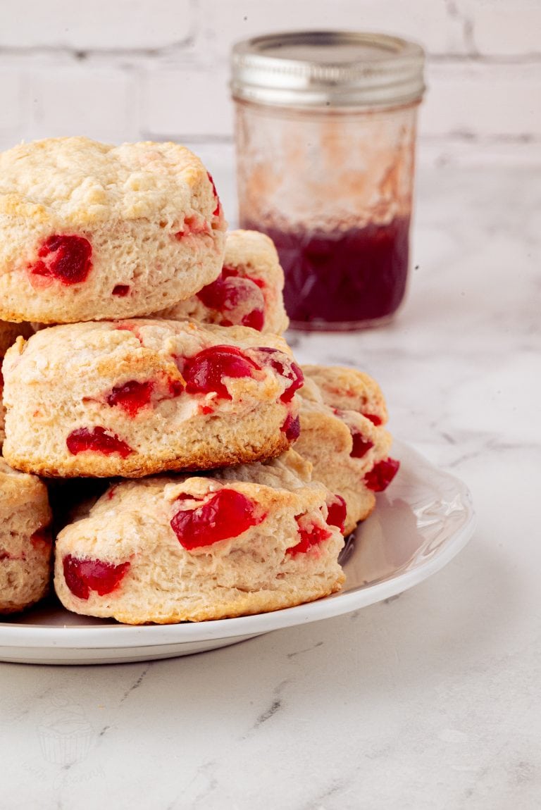 A stack of cherry scones, showcasing their delightful recipe with visible fruit pieces, sits on a white plate. In the background, a glass jar brimming with red jam complements the scene on a sleek marble countertop against a partially visible brick wall.