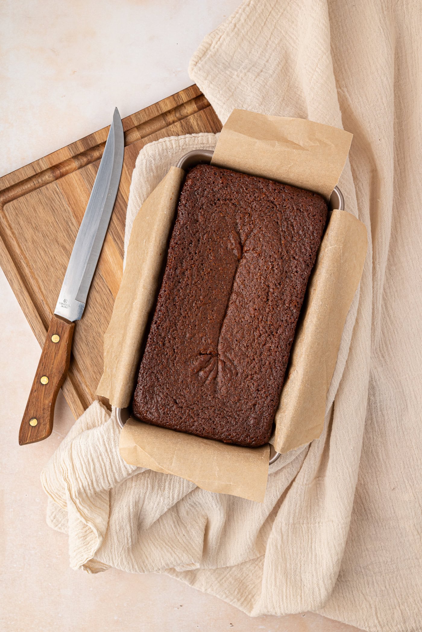 A freshly baked gingerbread cake, sits in a rectangular loaf tin lined with parchment paper on a wooden cutting board. A knife with a wooden handle rests beside it on a light fabric surface.