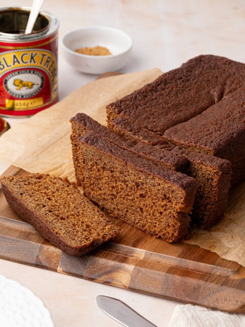 Sliced gingerbread loaf rests on a wooden cutting board, its deep brown hues inviting. Nearby, a can of black treacle and a small white dish with brown sugar add to the scene, accompanied by a knife and plate. The loaf is carefully placed on parchment paper.