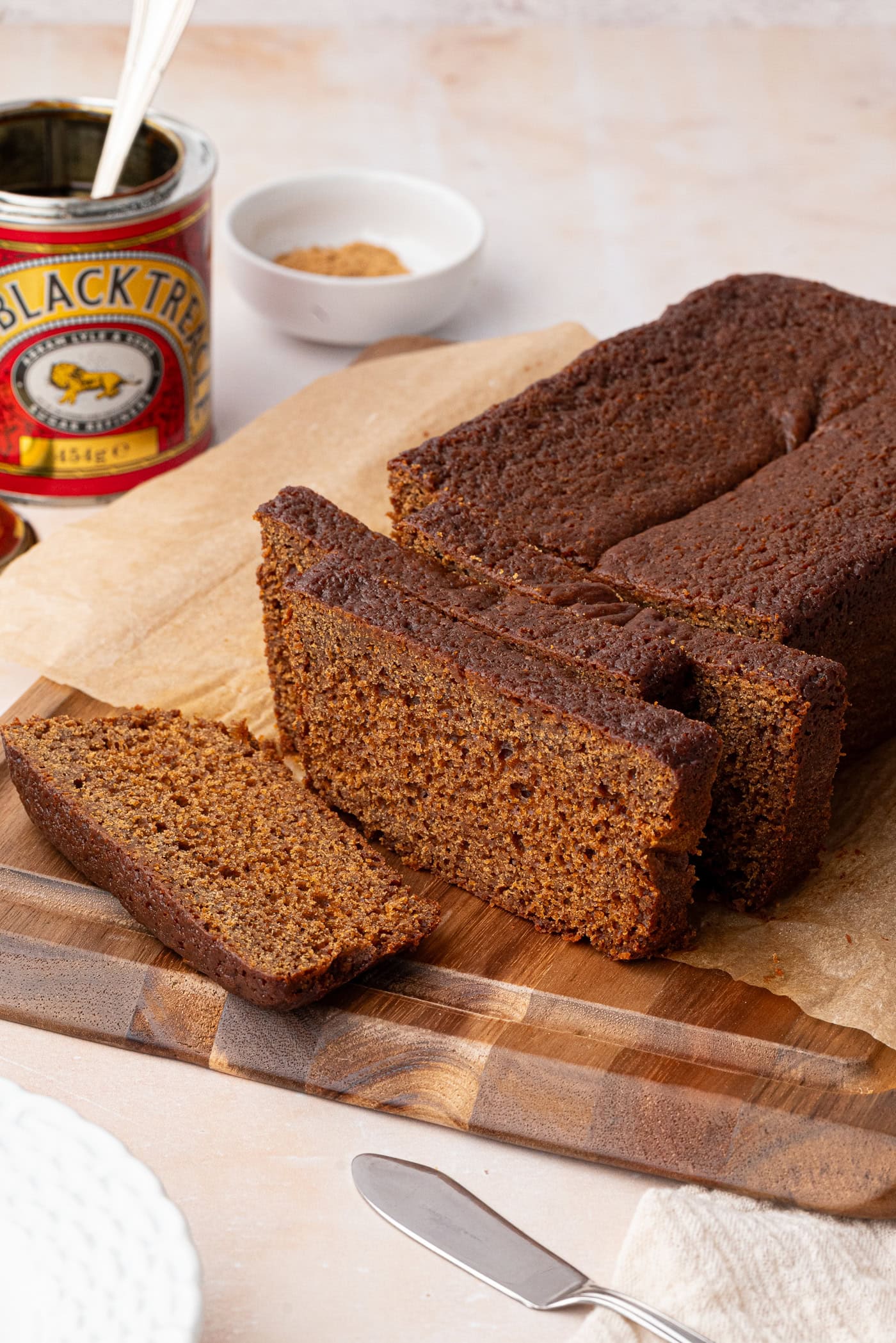 Sliced gingerbread loaf rests on a wooden cutting board, its deep brown hues inviting. Nearby, a can of black treacle and a small white dish with brown sugar add to the scene, accompanied by a knife and plate. The loaf is carefully placed on parchment paper.