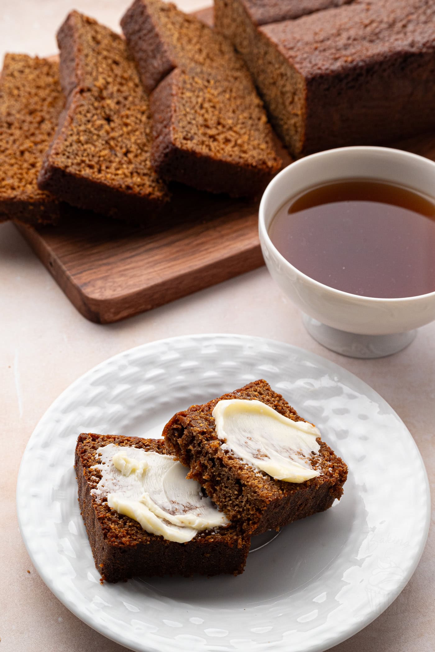 Slices of dark brown gingerbread loaf rest on a white plate with two pieces spread with butter. A cup of tea sits beside the plate, while more sliced gingerbread cake awaits on a wooden board in the background.