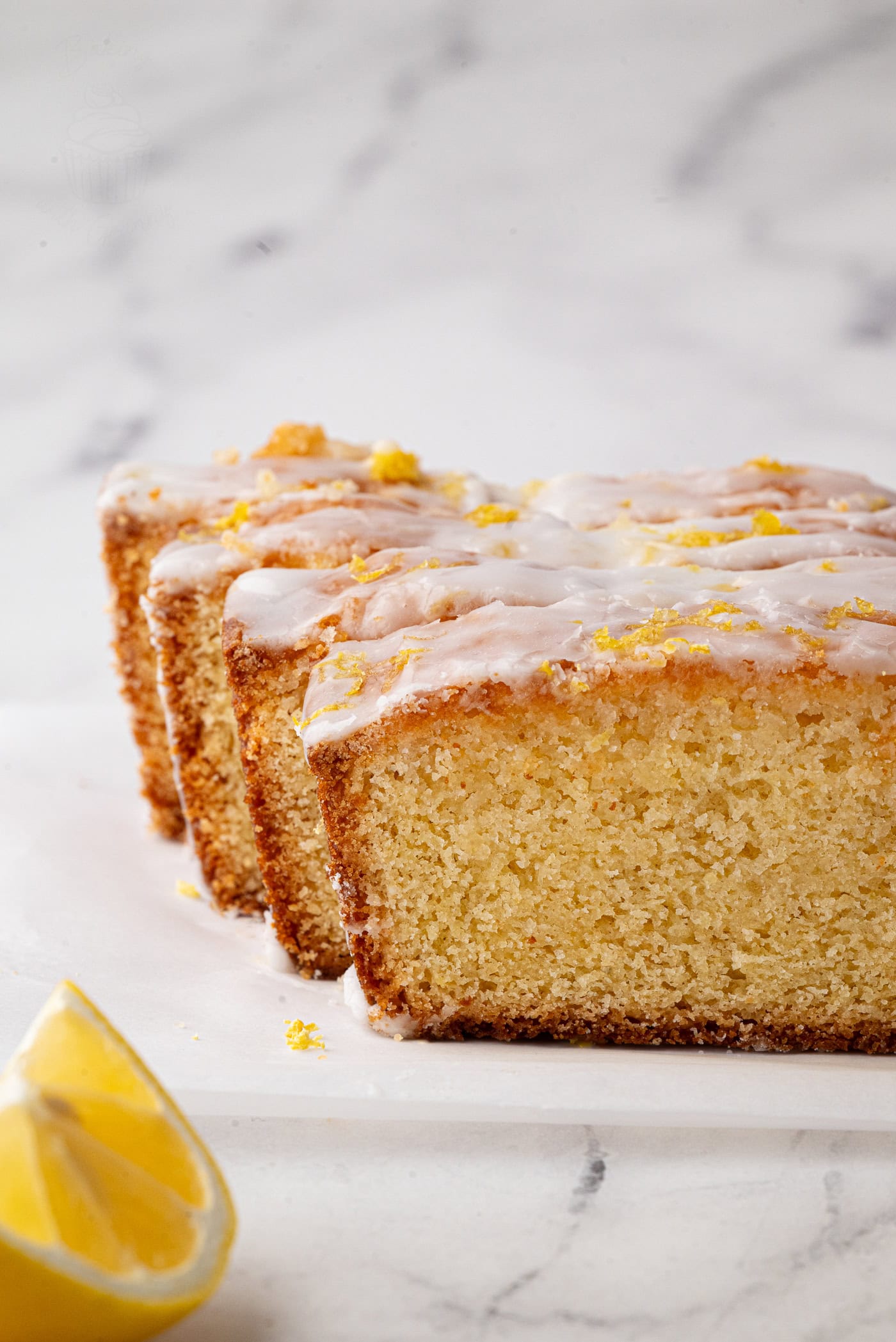 Slices of lemon drizzle cake topped with icing and lemon zest are neatly arranged on a white marble surface. A juicy wedge is placed in the foreground, amplifying the cakes citrus theme.