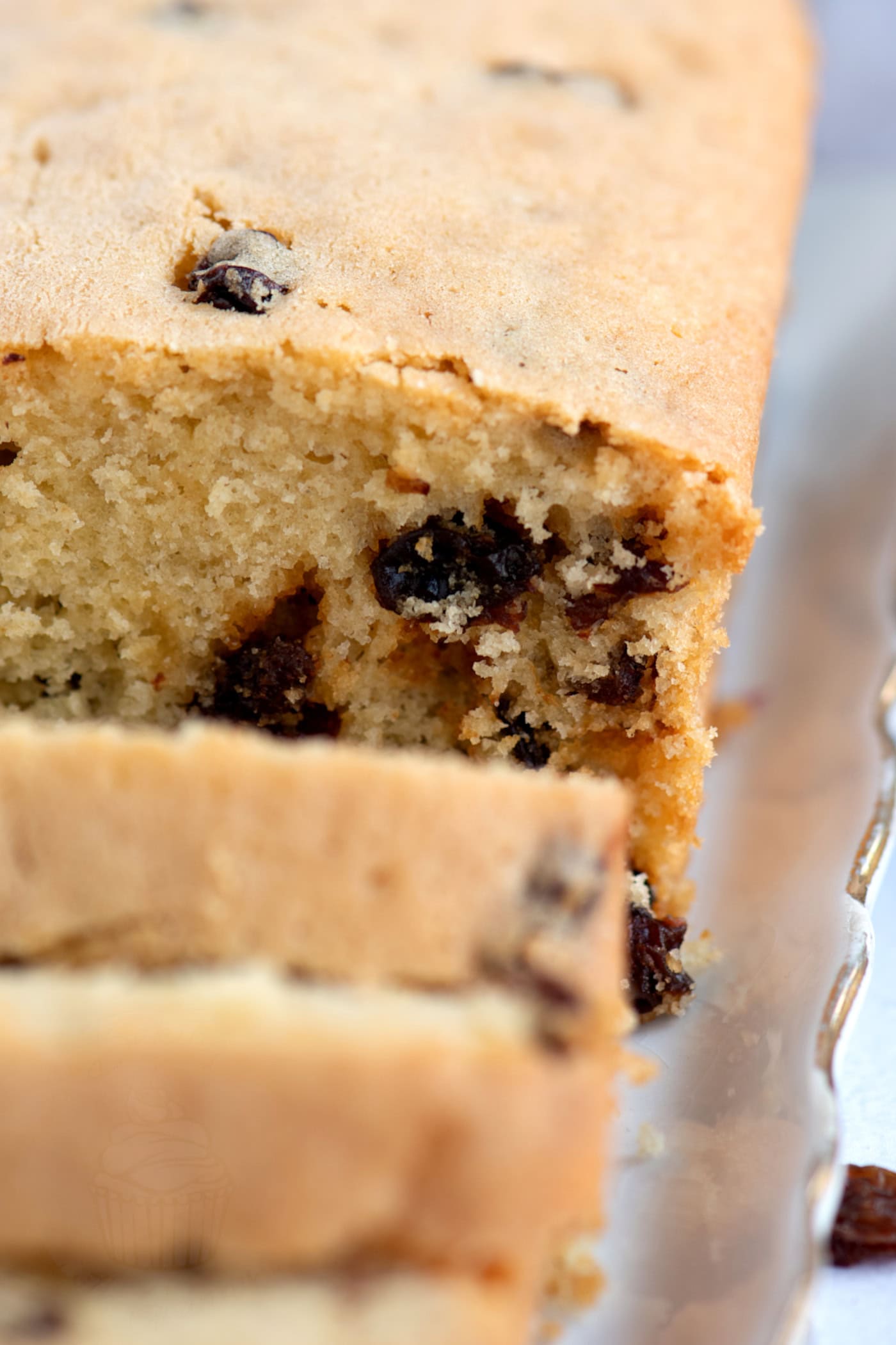 Close-up of a sliced loaf of light brown sultana cake on a white plate with gold trim. The cake contains visible pieces of sultanas, with a soft, crumbly texture. Two slices have been cut from the loaf, revealing the moist interior.