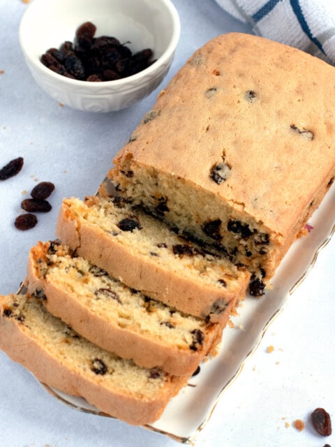 A loaf of Scottish sultana cake recipe, sits partially sliced on a plate, accompanied by a small bowl of sultanas. In the background, a checked cloth rests on the light surface.