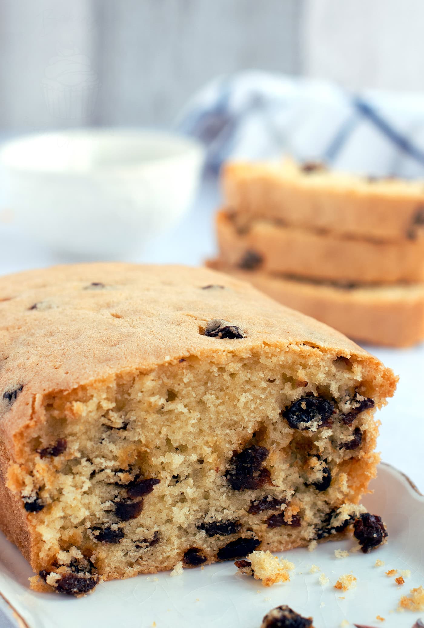 A close-up of a sliced loaf of sultana cake on a plate. The cake is moist with visible sultanas. In the background, more slices are stacked with a napkin and bowl slightly blurred.