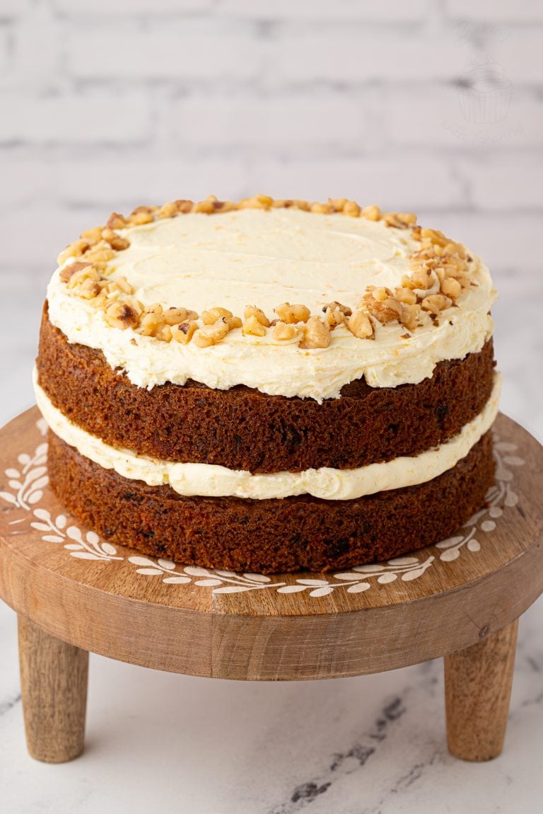 Two-layer carrot cake topped with buttercream and chopped walnuts, displayed on a wooden cake stand with inlaid detail.