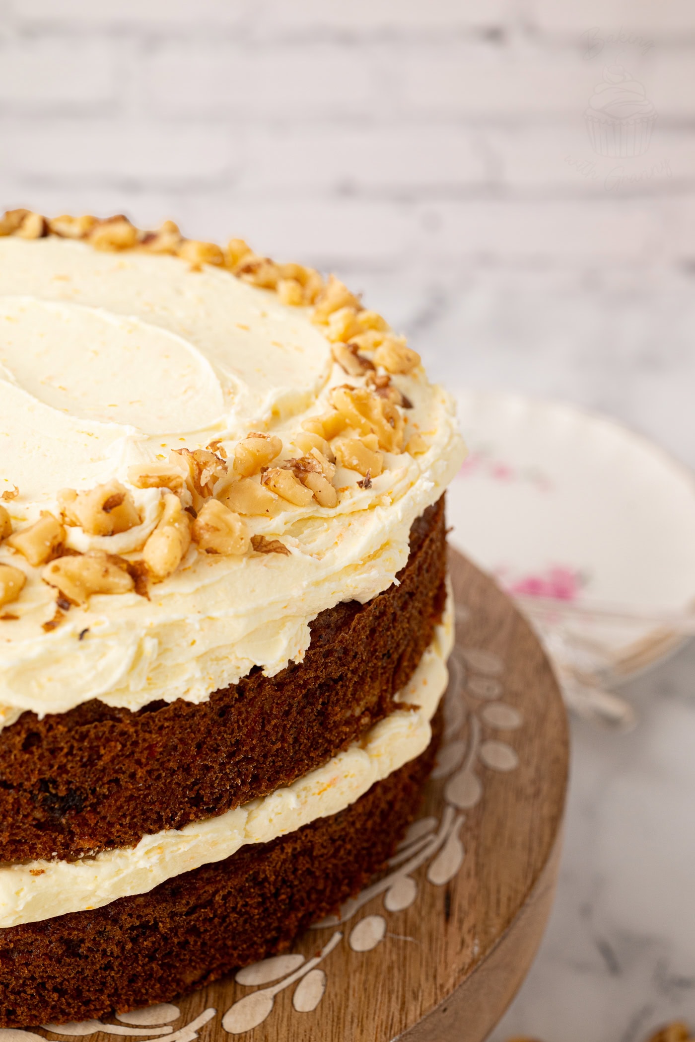 Close-up of the top edge of a carrot cake, showing creamy frosting swirls and a ring of chopped walnuts on a rustic cake stand.