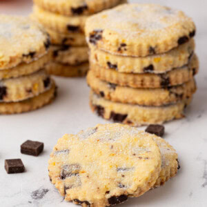 Focus on a single sugared chocolate orange shortbread biscuit, with several stacks of biscuits and an orange half in soft focus behind.