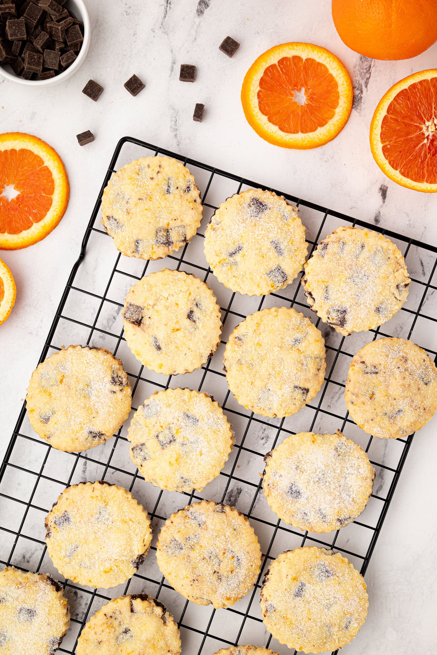 Overhead view of chocolate orange shortbread cookies on a wire rack, surrounded by orange slices and scattered chocolate chunks.