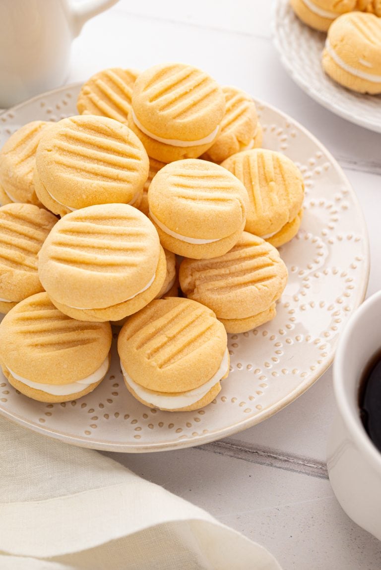 Overhead view of custard cream biscuits arranged on a cream plate beside white coffee mugs and napkins.