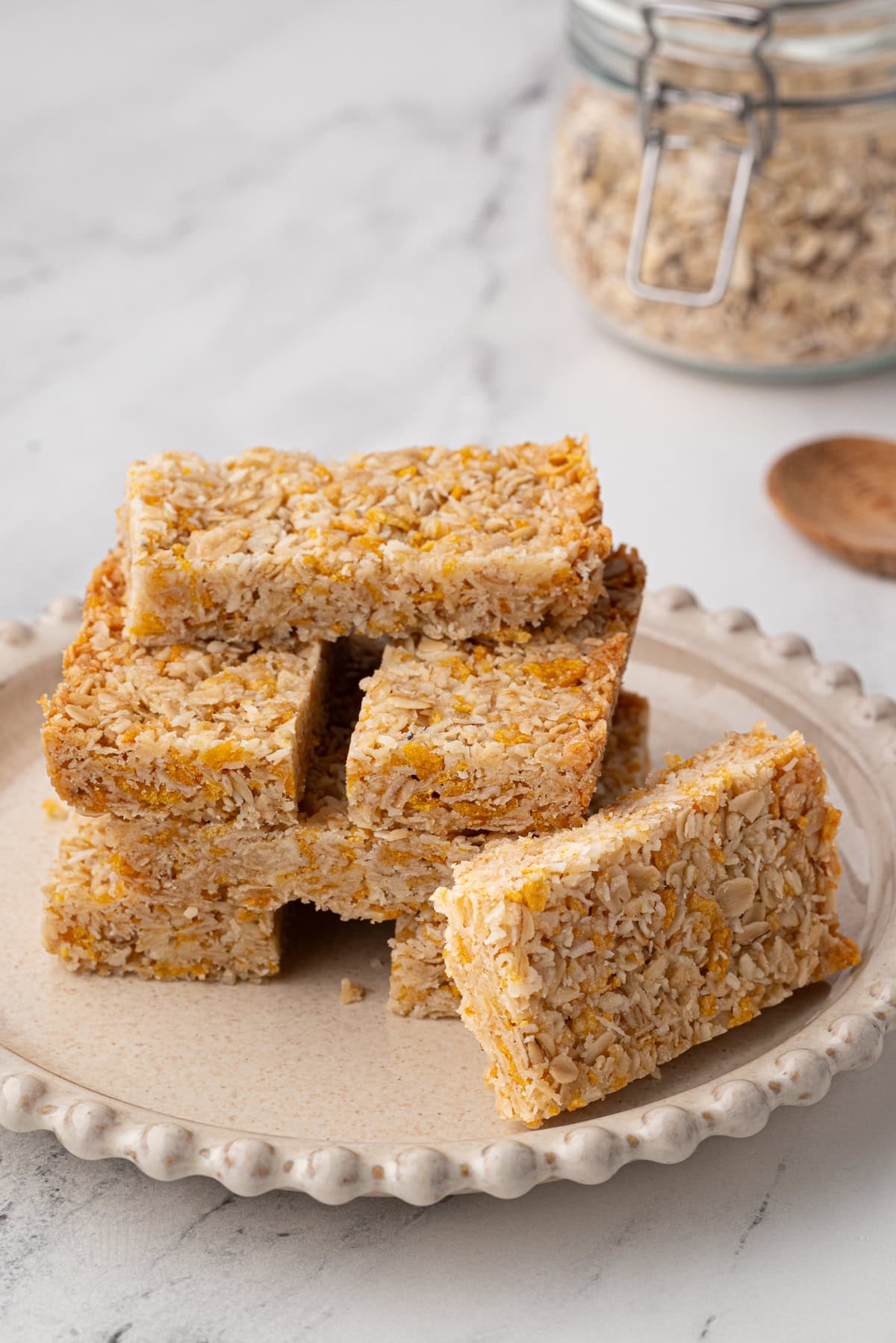 Stack of honey slice bars on a decorative ceramic plate, showing the golden oaty texture with flecks of cornflakes and coconut.