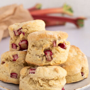 Plate of stacked Rhubarb Scones with visible rhubarb pieces, and raw rhubarb stalks visible in the background.