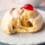 Side view of a Belgian bun with a bite taken out, topped with white icing and a red glacé cherry, served with tea.