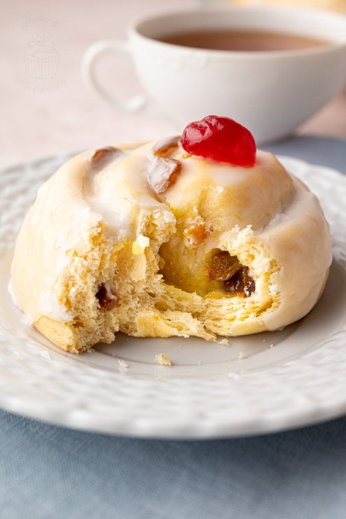 Side view of a Belgian bun with a bite taken out, topped with white icing and a red glacé cherry, served with tea.