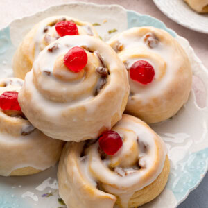 Stack of glazed Belgian buns with red cherries on top, served on a patterned plate, ideal for British teatime.