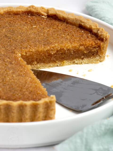 A slice of traditional treacle tart being served from a white dish. The sticky, golden filling contrasts with the buttery pastry base.