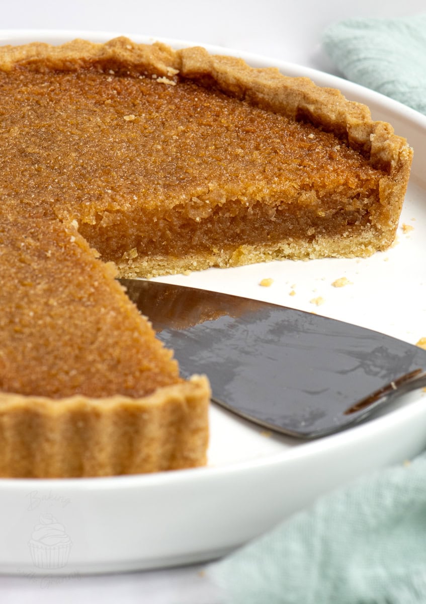 A slice of traditional treacle tart being served from a white dish. The sticky, golden filling contrasts with the buttery pastry base.
