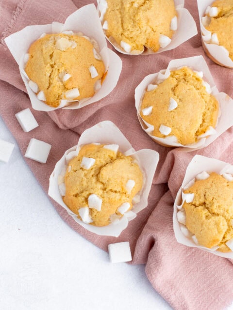 Freshly baked Sair Heidies fairy cakes in tulip muffin cases, decorated with broken sugar cubes on top, sitting on a pink cloth.