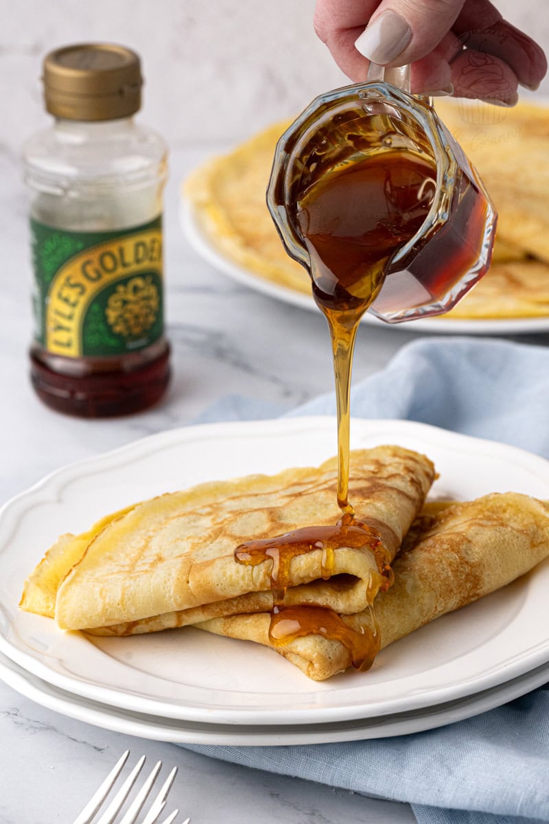Golden syrup being poured over folded traditional British pancakes on a white plate.