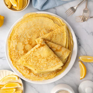 Overhead view of a stack of thin crepes with lemon wedges and sugar for Pancake Day.