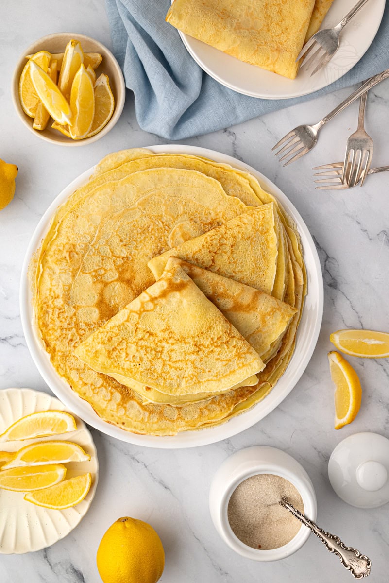 An overhead view (flat lay) showing a stack of thin, large round British pancakes on a white plate, surrounded by bowls of lemon slices, a sugar pot, and two silver forks.