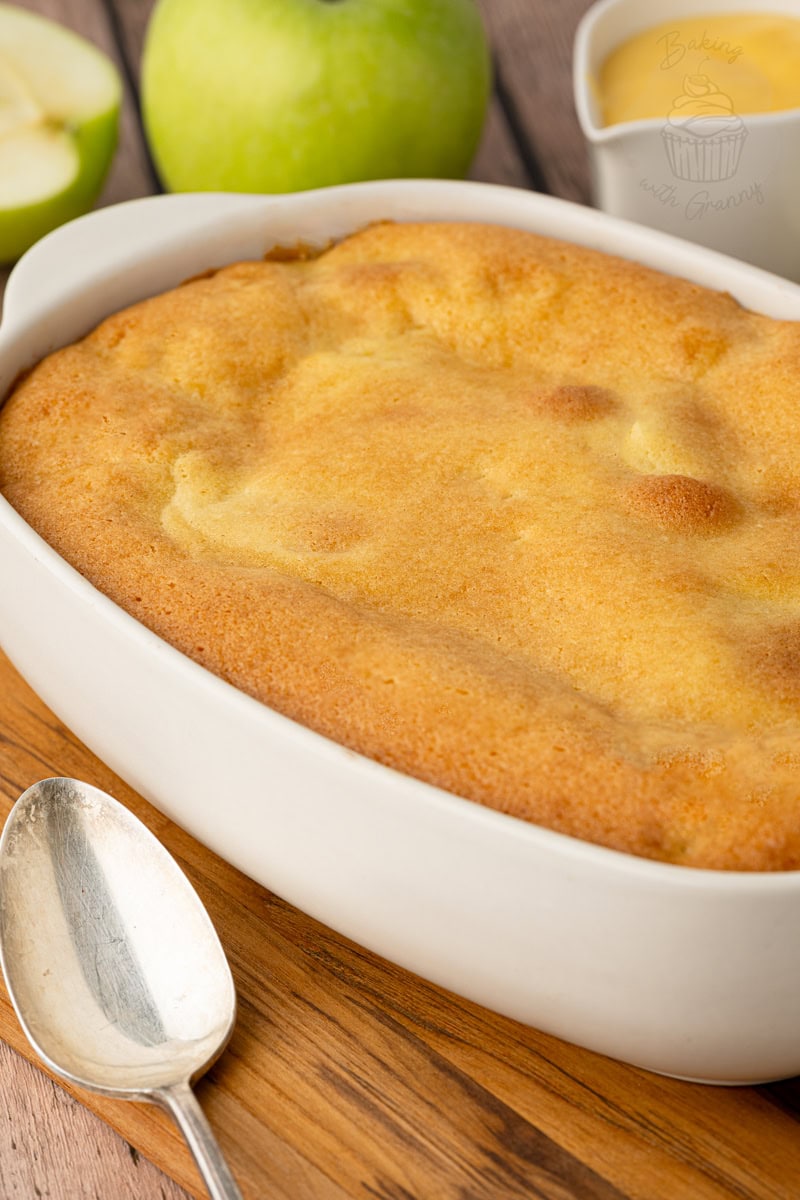 A freshly baked old-fashioned apple sponge pudding with a golden-brown crust, shown next to a silver spoon and whole green Bramley apples.