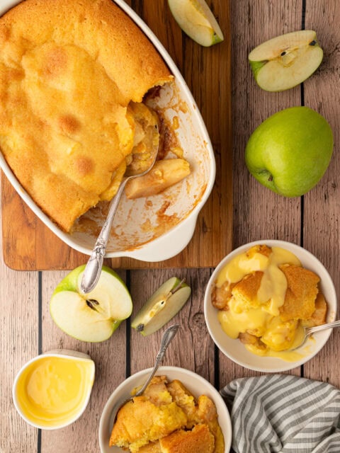 An old-fashioned British Eve's pudding dessert served in a bowl and covered in smooth yellow custard, styled on a rustic wooden background.