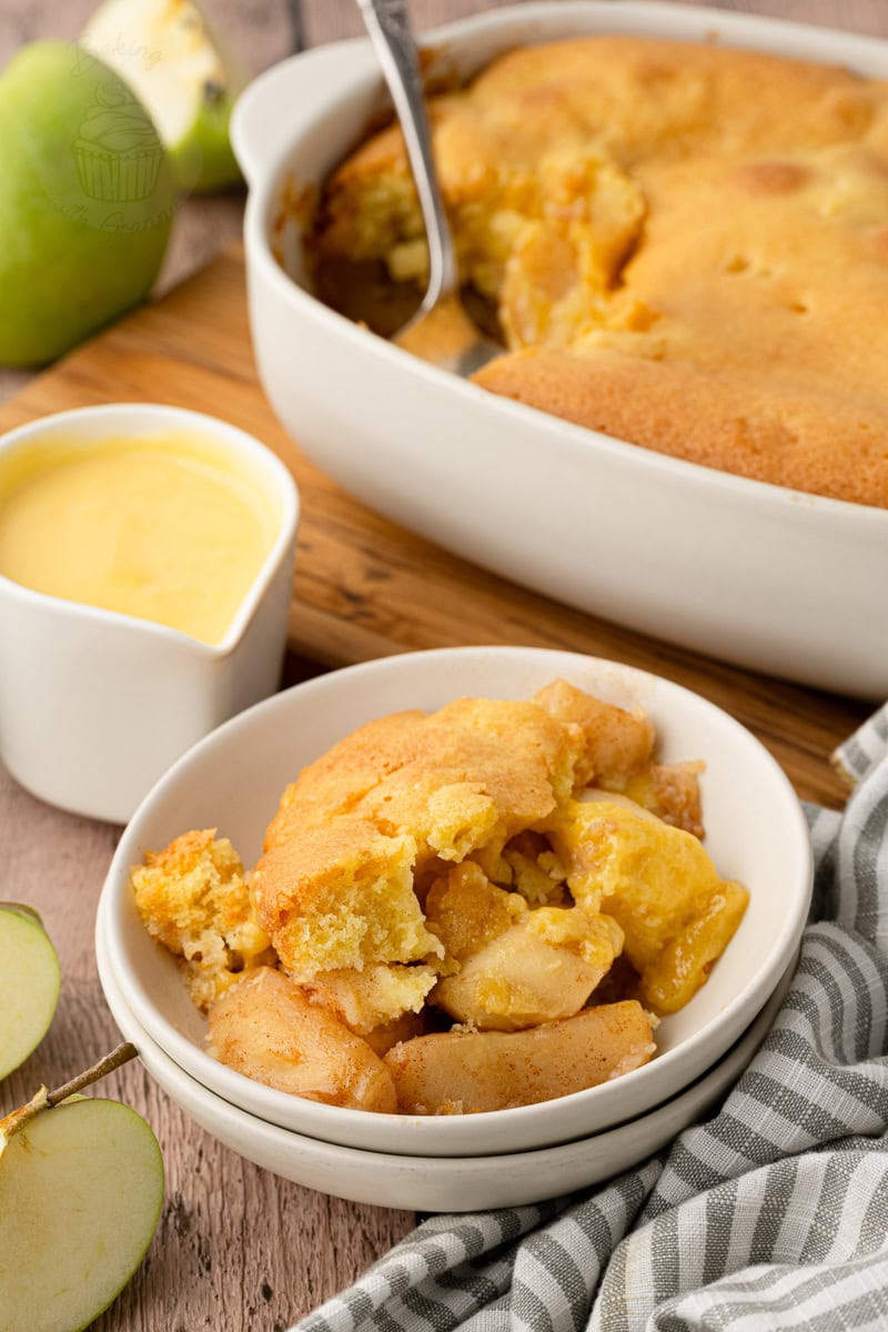 A single serving of easy apple sponge pudding in a small bowl, positioned next to a white jug of custard and the main baking dish.