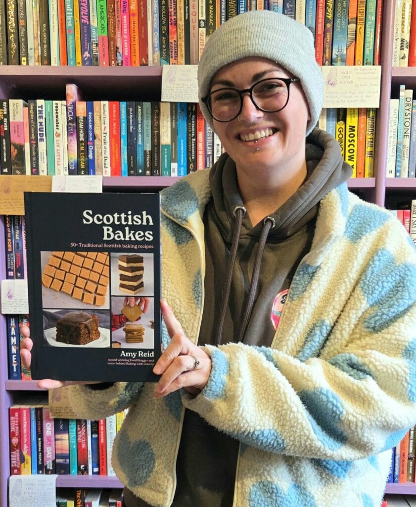 Amy Beth Reid, author of Scottish bakes, holding a copy of her debut cookbook against a book shop backdrop.