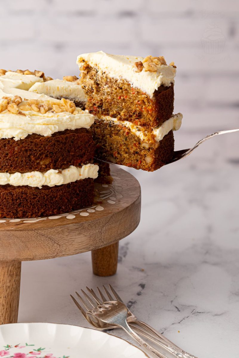 Carrot Cake A slice of carrot cake with walnuts and frosting being lifted from a whole cake on a wooden stand, ready to serve.