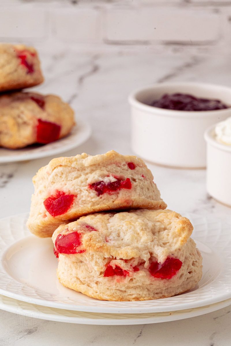 Cherry Scones A duo of cherry scones sits enticingly atop a white plate, bursting with juicy red cherries. In the background, additional cherry scones are accompanied by small bowls of jam and cream on a sleek marble surface.