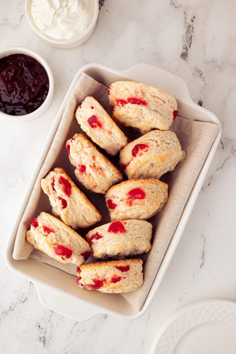 Cherry Scones A rectangular dish holds several easy cherry scones with vibrant red glace cherries pieces. The dish is lined with parchment paper. Nearby, small bowls are filled with cream and dark red jam, all elegantly arranged on a light-coloured marble surface.