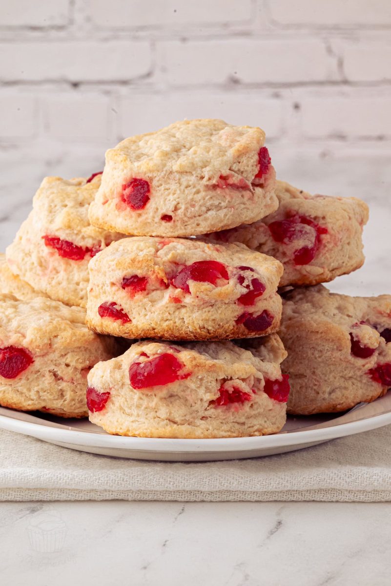 Cherry Scones A plate of freshly baked cherry scones featuring bits of bright red glace cherries, stacked in a pyramid shape. The scones have a golden-brown crust and are set against a white brick background, offering an irresistible cherry scones recipe visual delight.