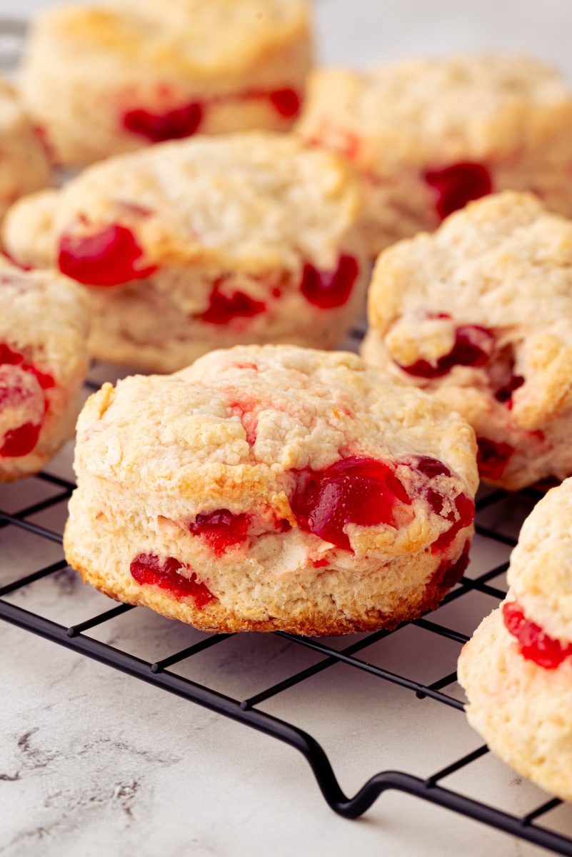 Cherry Scones Close-up of cherry scones with visible chunks of glace cherries, placed on a black wire cooling rack. The scones have a golden, crumbly texture, and the cherries add a pop of red colour. The background is a light, textured surface.