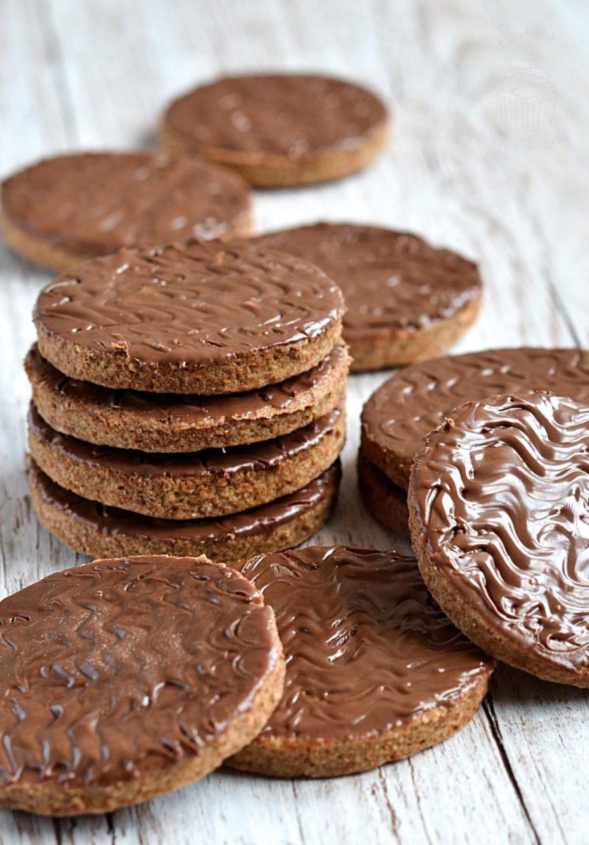 Chocolate Digestive Stacked chocolate digestive cookies surrounded by more on a whitewashed wooden table, showing their glossy chocolate tops and golden brown biscuit base.