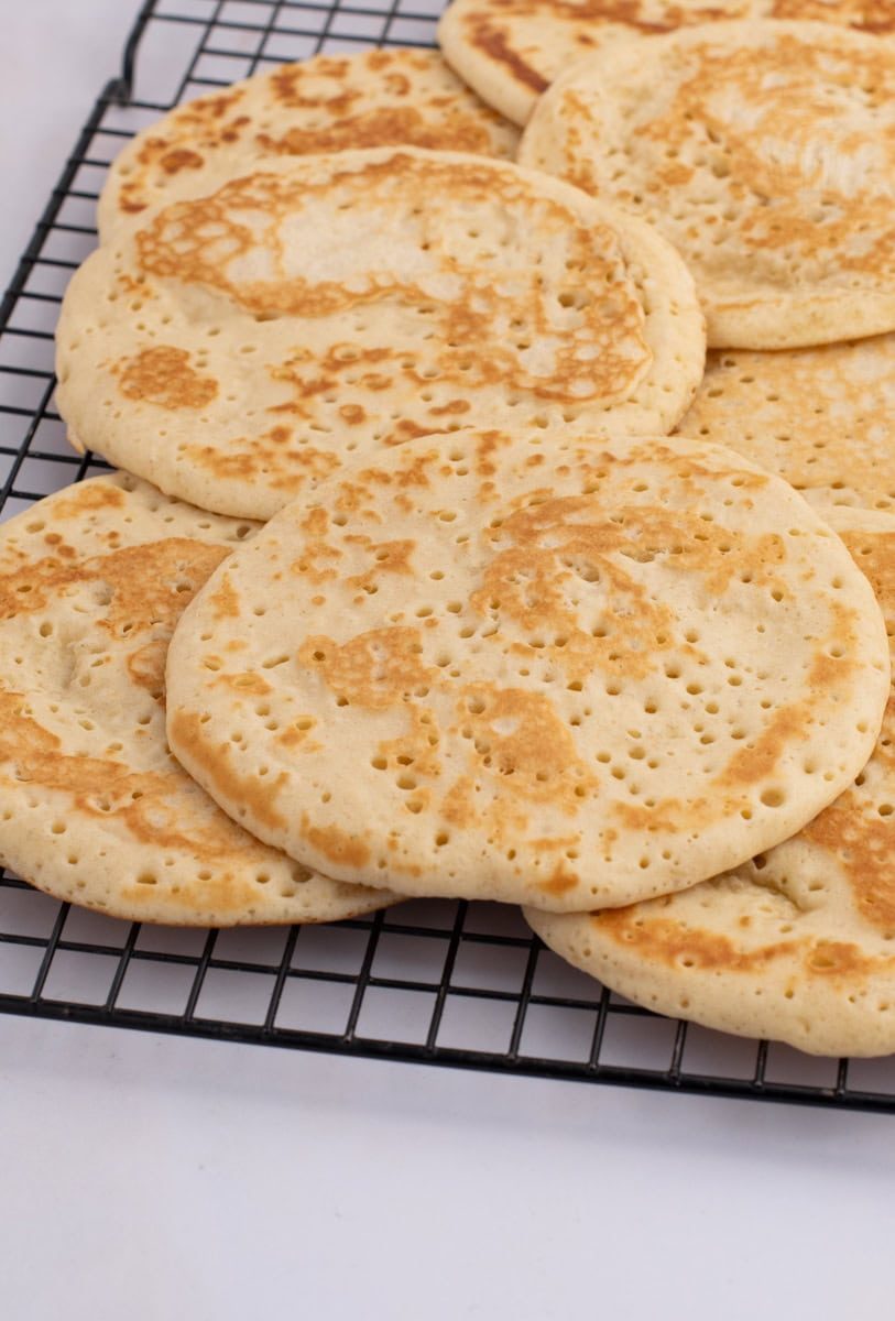 Scottish Crumpets A stack of homemade Scottish crumpets cools on a black wire rack. These golden brown delights, with their characteristic small holes, perfectly capture the light and airy texture that makes this recipe a beloved classic.