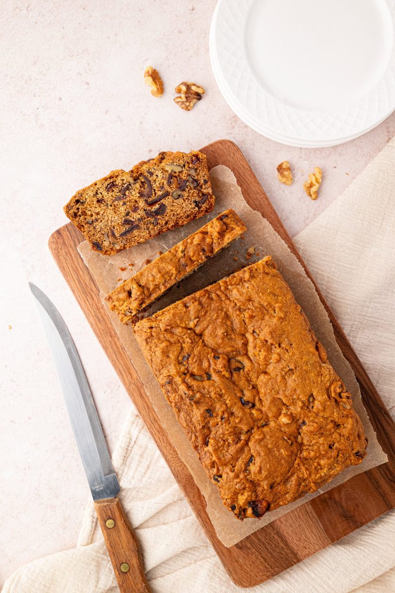 A date and walnut loaf rests on a wooden cutting board. Two slices have been cut, with one placed beside the loaf. A knife lies close, while a white plate and scattered walnuts add to the inviting scene in the background.