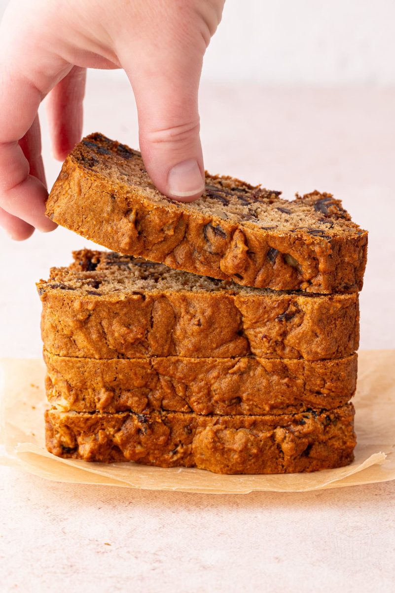 A hand lifts a slice of date and walnut loaf from a stack of four on parchment paper, revealing its golden-brown crust dotted with walnut and date pieces. This delicious treat is reminiscent of a classic date and walnut loaf recipe. The softly blurred background enhances its allure.