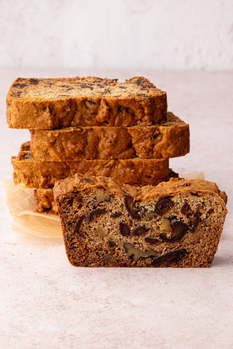 A sliced date and walnut loaf reveals its moist, dense interior, adorned with visible walnuts and dates on a light surface. In the background, several golden-brown slices are stacked with parchment paper, showcasing an irresistible take on a classic recipe.