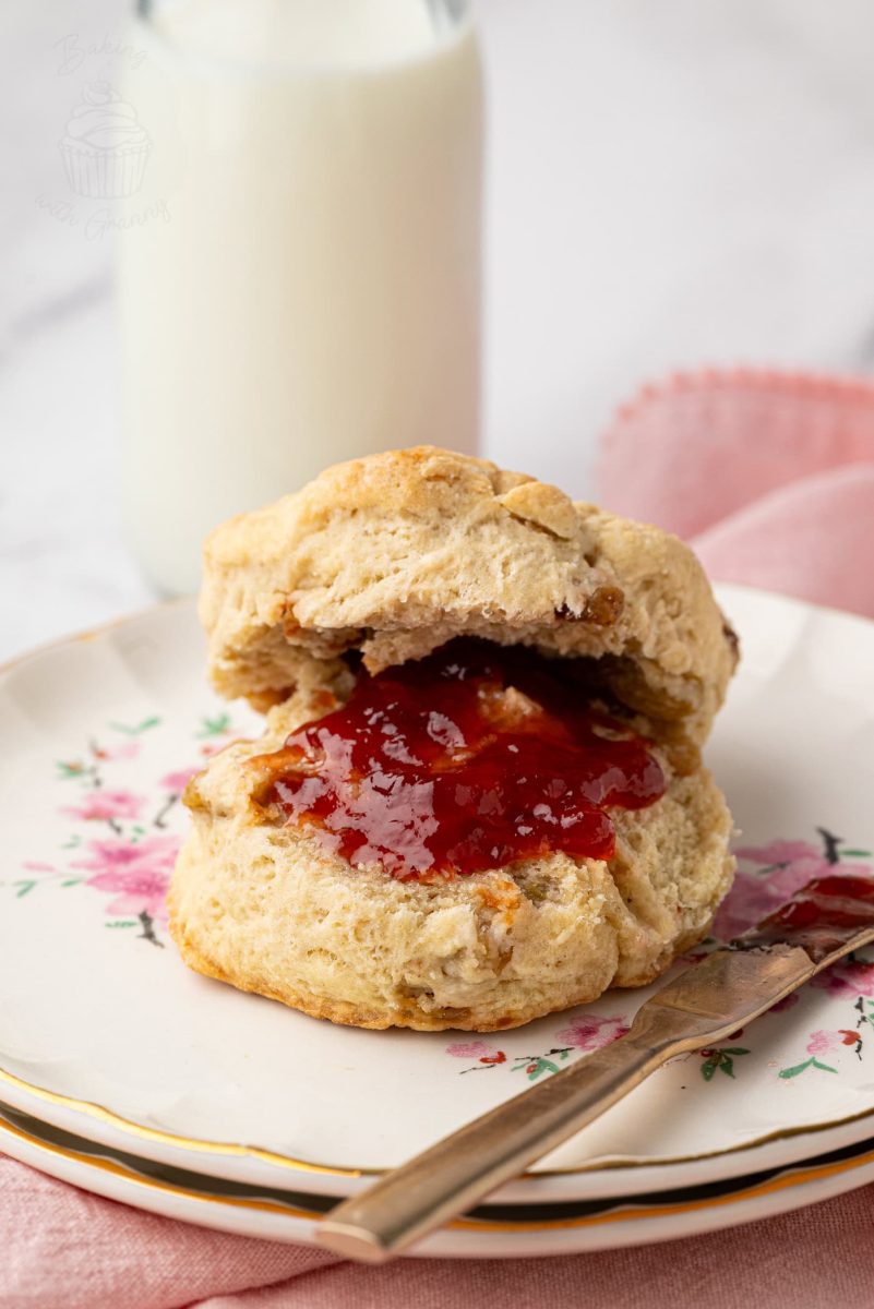 Fruit Scones Close-up of a golden fruit scone filled with sultanas, spread with strawberry jam, next to a glass of milk.