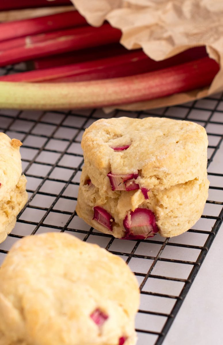Rhubarb Scones Individual Rhubarb Scone on a cooling rack in front of fresh seasonal rhubarb stems, within a brown paper bag.