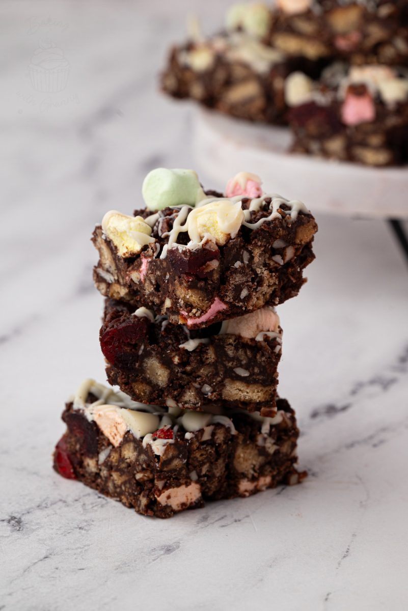 Rocky Road Three stacked rocky road squares, complete with visible nuts, marshmallows, and chocolate, are drizzled with white chocolate. They sit on a marble surface like a visual invitation from your favourite rocky road recipe, with more squares blurred in the background.