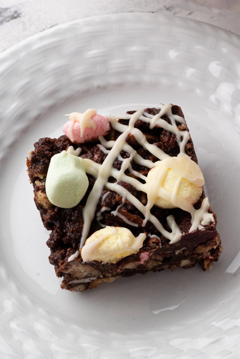 Rocky Road A square slice of rocky road tray bake rests on a white plate, adorned with colourful marshmallows and drizzled with white chocolate in a crisscross pattern.