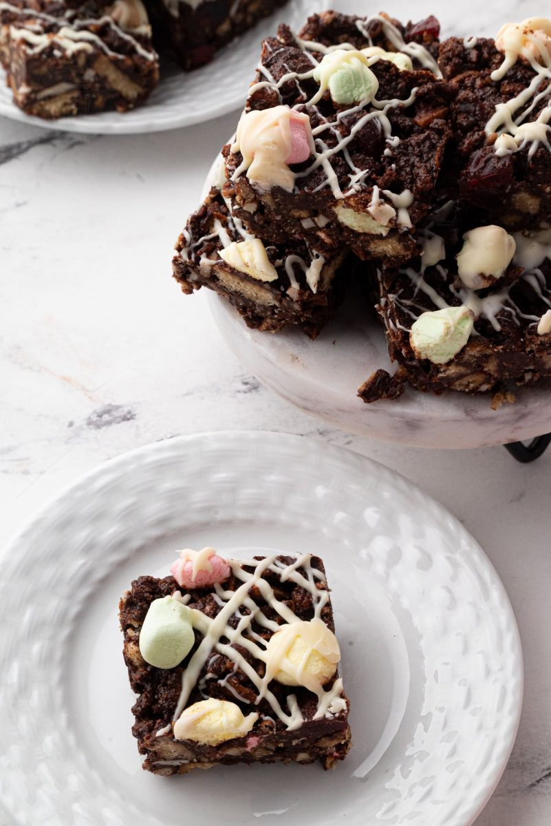 Rocky Road A plate of delightful rocky road squares is topped with colourful marshmallows and a luscious white chocolate drizzle. In the background, another plate offers more of these tempting rocky road treats, all beautifully arranged on a light marble surface.