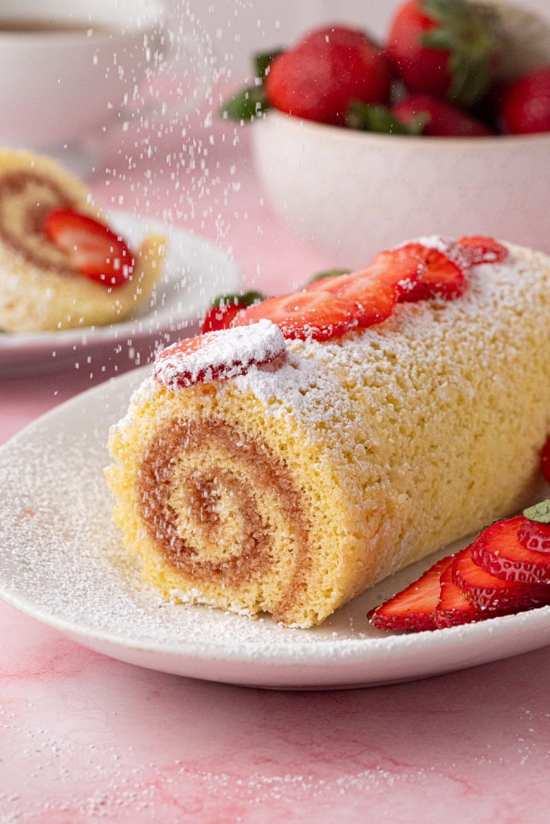 Swiss Roll A sliced Swiss roll cake, crafted from a classic Swiss roll recipe, is topped with icing sugar and fresh strawberry slices on a white plate. A bowl of strawberries graces the background on a light pink surface.