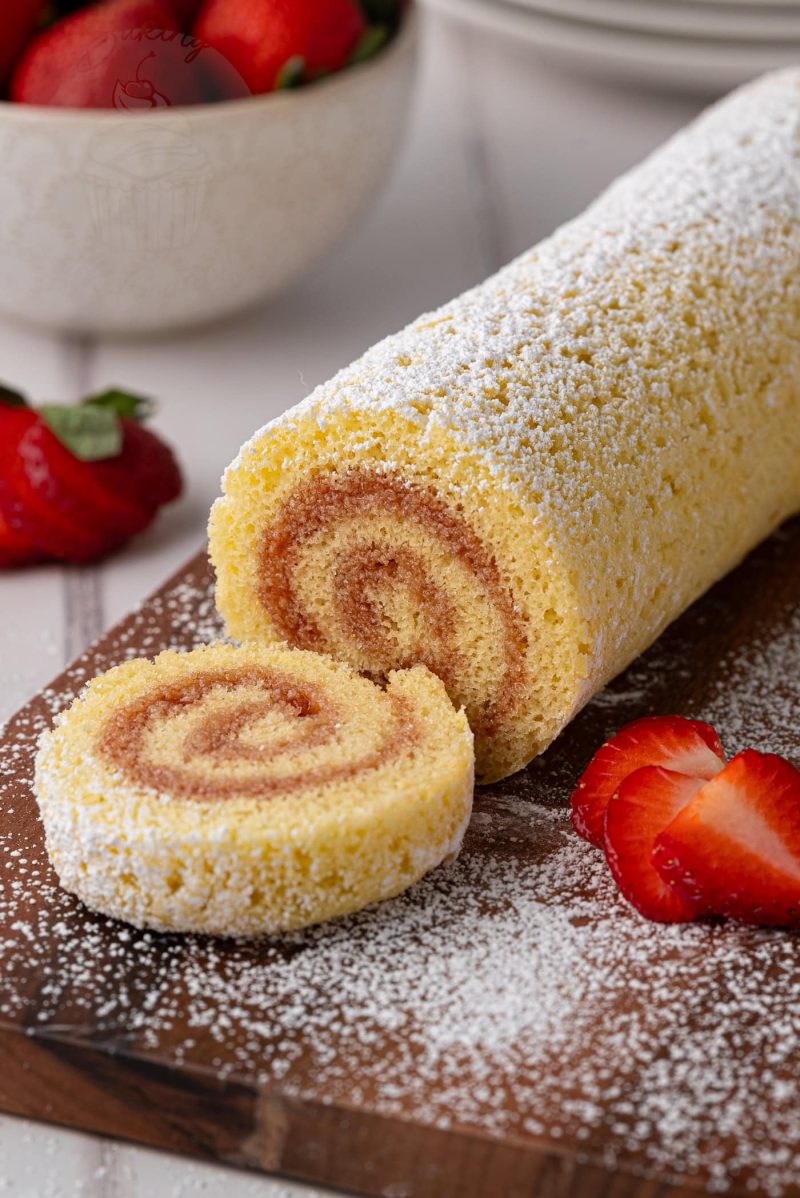 Swiss Roll A rolled sponge cake, reminiscent of a classic Swiss Roll recipe, is dusted with icing sugar and sliced on a wooden board. Fresh strawberries are placed around it, with a bowl of strawberries in the background.