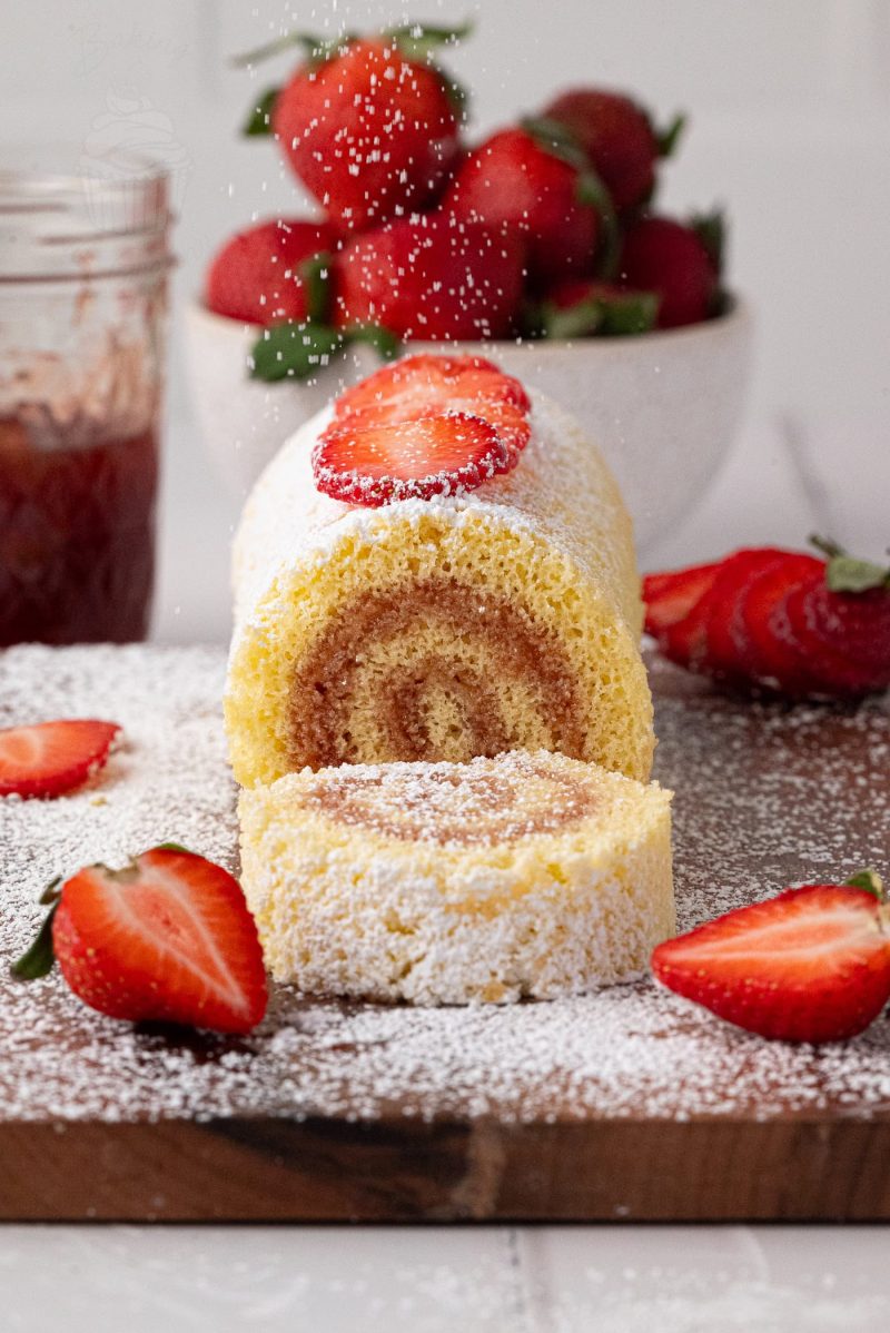 Swiss Roll A simple Swiss roll recipe comes to life with a cake dusted in icing sugar and surrounded by sliced strawberries on a wooden board. In the background, a bowl of whole strawberries and a jar of jam complete the enticing scene.