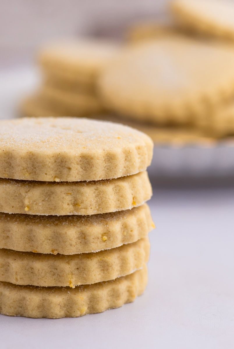A stack of five round Tantallon Cakes with scalloped edges is in focus. The biscuits are light golden brown with a dusting of sugar on top, reminiscent of classic shortbread. More cookies are blurred in the background on a white surface.