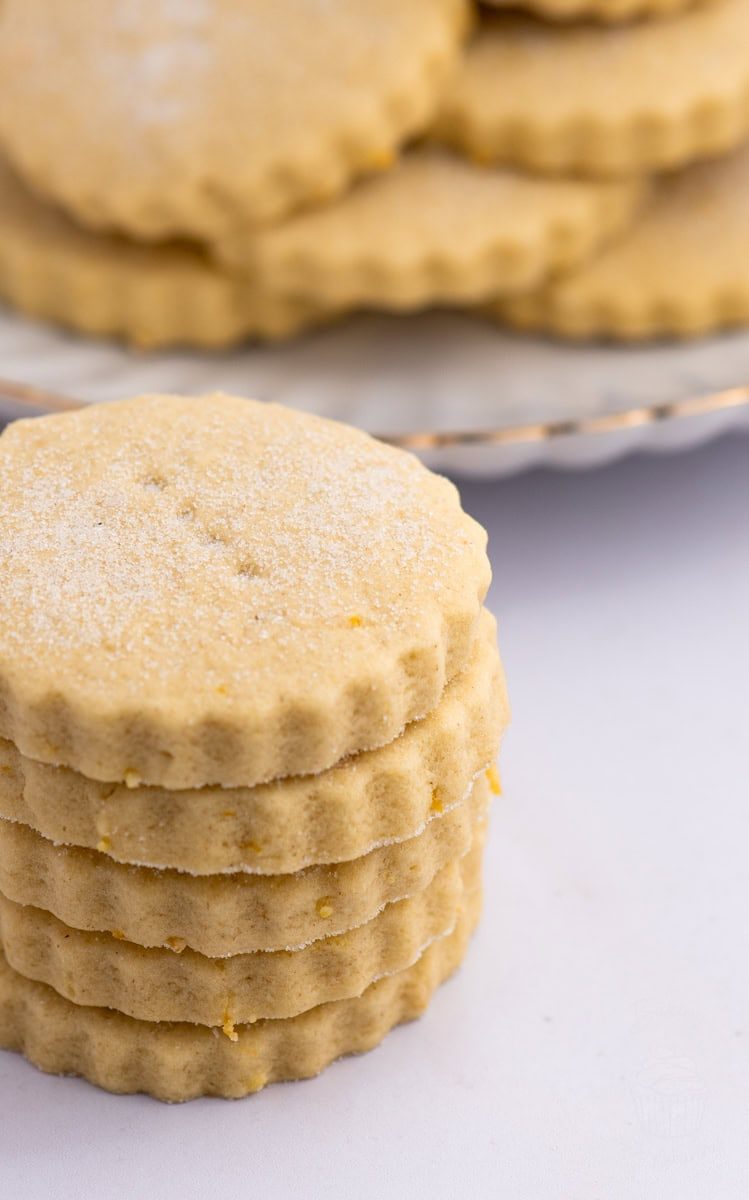 A stack of five round, scalloped-edged, light brown biscuits with a sugar coating and specks of lemon rind, evokes the essence of a classic recipe for Tantallon Cakes. More cookies are blurred in the background on a plate. The cookies appear soft and crumbly, set against a white surface.