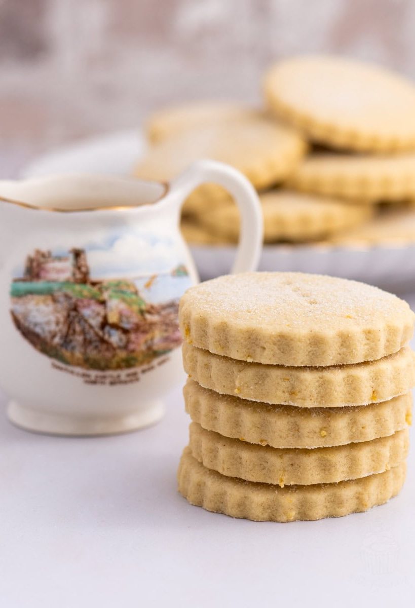 A stack of six round, scalloped-edge Tantallon cakes sits in the foreground. Behind them is a decorative jug with a scenic illustration of Tantallon Castle in North Berwick. This delightful scene could inspire your next baking recipe, with more biscuits visible in the blurred background.
